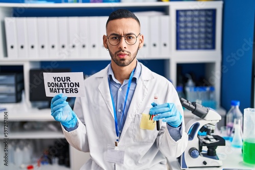 Young hispanic man working at scientist laboratory holding your donation matters holding blood sample skeptic and nervous, frowning upset because of problem. negative person.
