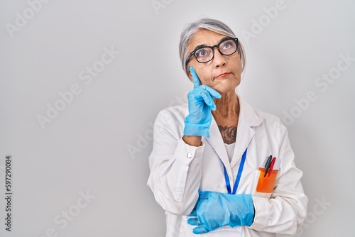 Middle age woman with grey hair wearing scientist robe with hand on chin thinking about question  pensive expression. smiling with thoughtful face. doubt concept.