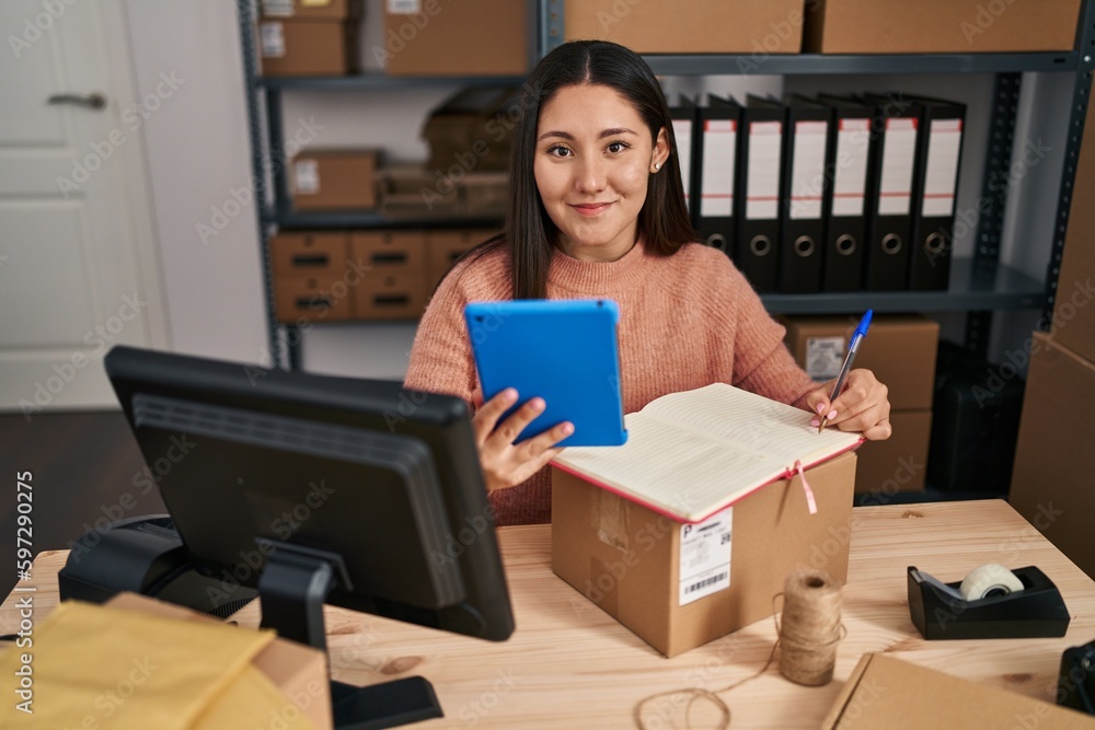 Young hispanic woman ecommerce business worker writing on notebook and using touchpad at office Young hispanic woman ecommerce business worker writing on notebook and using touchpad at office