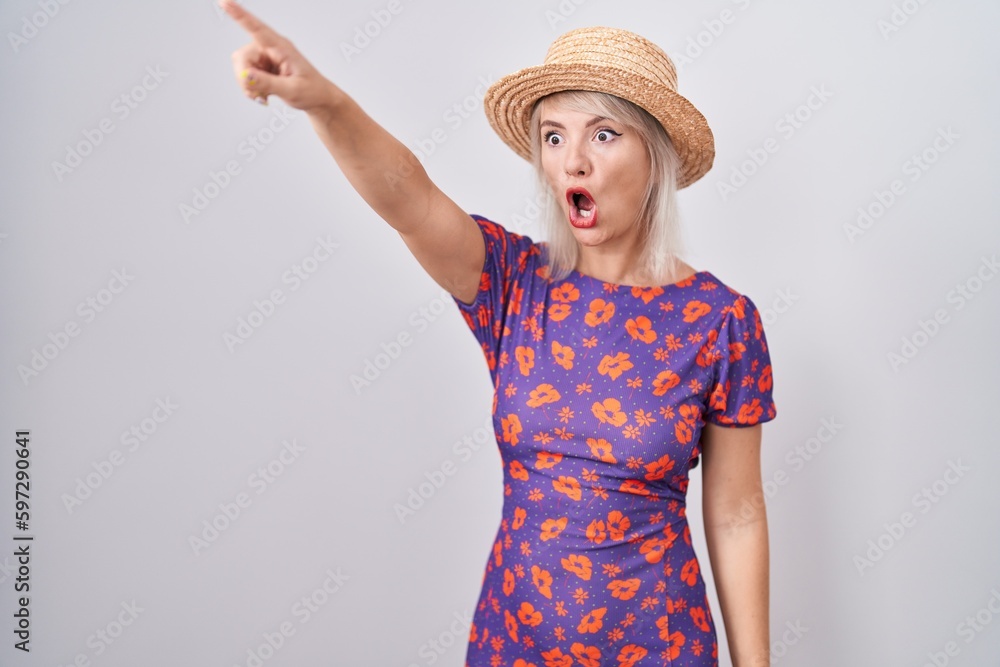 Young caucasian woman wearing flowers dress and summer hat pointing with finger surprised ahead, open mouth amazed expression, something on the front Young caucasian woman wearing flowers dress and summer hat pointing with finger surprised ahead, open mouth amazed expression, something on the front