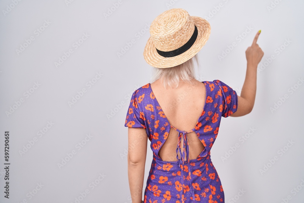 Young caucasian woman wearing flowers dress and summer hat posing backwards pointing ahead with finger hand Young caucasian woman wearing flowers dress and summer hat posing backwards pointing ahead with finger hand