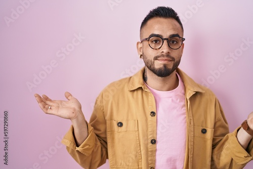 Young hispanic man standing over pink background clueless and confused expression with arms and hands raised. doubt concept.
