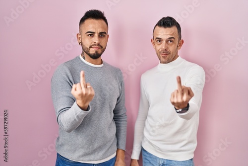 Homosexual couple standing over pink background showing middle finger, impolite and rude fuck off expression