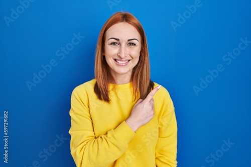 Young woman standing over blue background cheerful with a smile on face pointing with hand and finger up to the side with happy and natural expression
