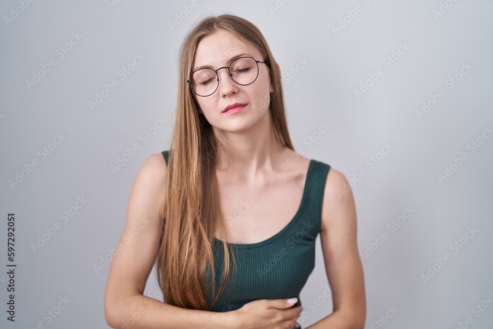 Young caucasian woman standing over white background with hand on stomach because indigestion, painful illness feeling unwell. ache concept. Young caucasian woman standing over white background with hand on stomach because indigestion, painful illness feeling unwell. ache concept.