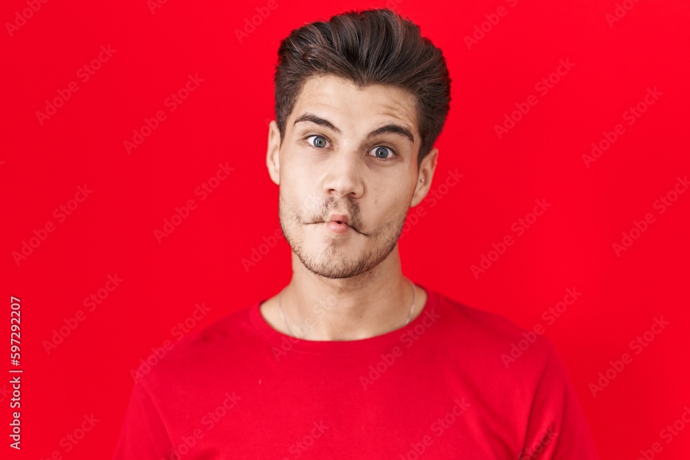Young hispanic man standing over red background making fish face with lips, crazy and comical gesture. funny expression.