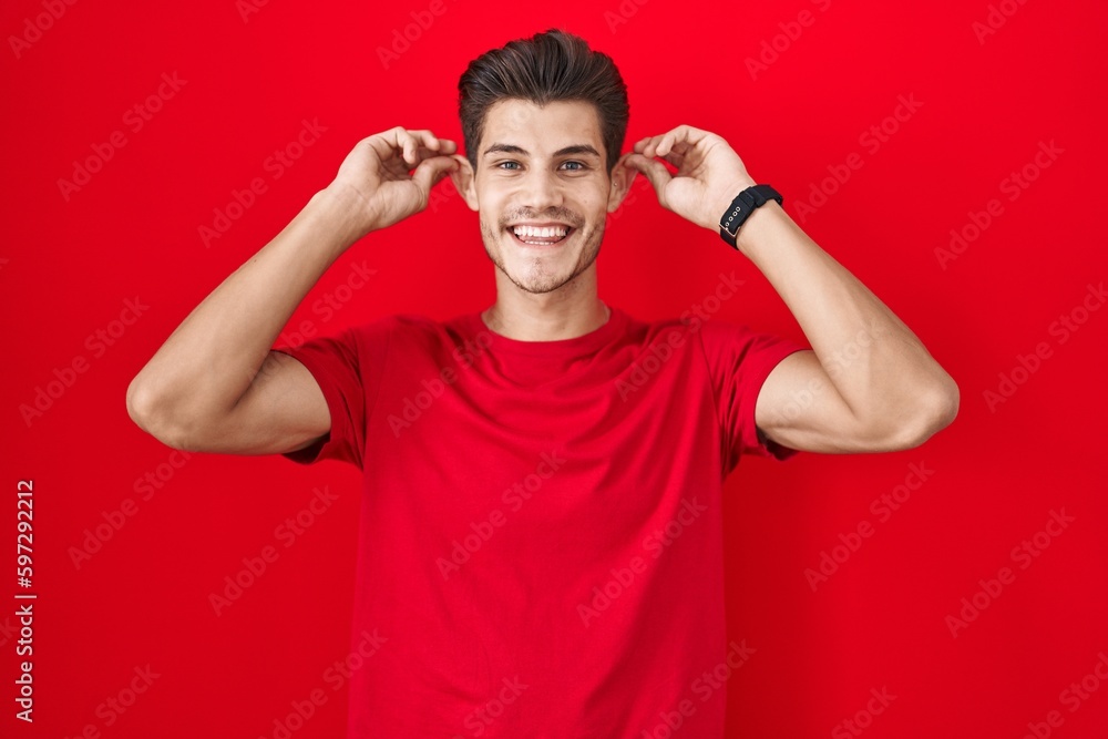 Young hispanic man standing over red background smiling pulling ears with fingers, funny gesture. audition problem Young hispanic man standing over red background smiling pulling ears with fingers, funny gesture. audition problem