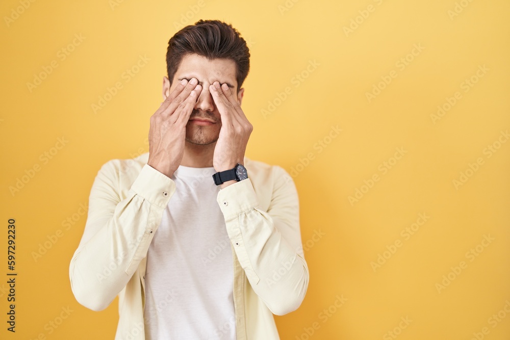 Young hispanic man standing over yellow background rubbing eyes for fatigue and headache, sleepy and tired expression. vision problem Young hispanic man standing over yellow background rubbing eyes for fatigue and headache, sleepy and tired expression. vision problem
