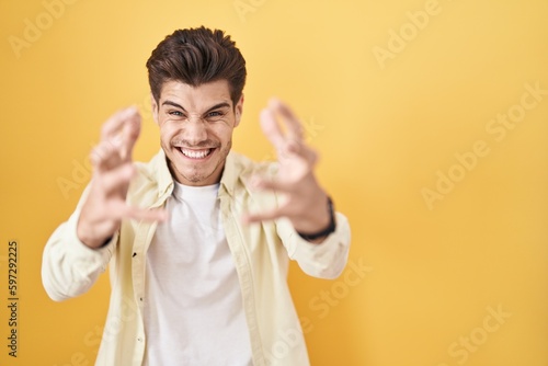 Young hispanic man standing over yellow background shouting frustrated with rage, hands trying to strangle, yelling mad