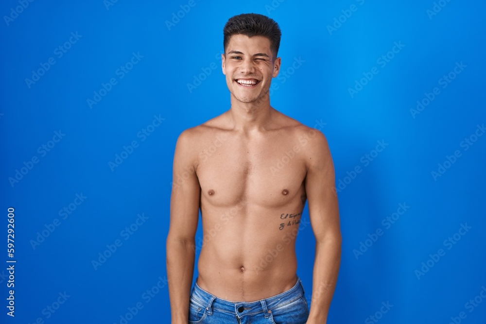 Young hispanic man standing shirtless over blue background winking looking at the camera with sexy expression, cheerful and happy face.