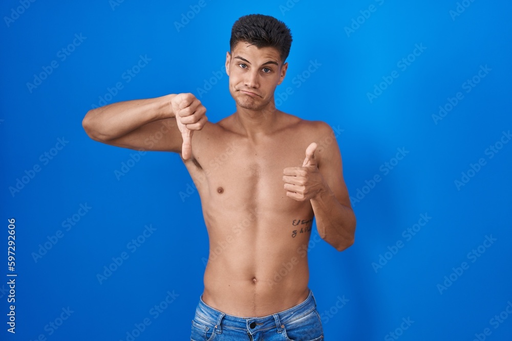 Young hispanic man standing shirtless over blue background doing thumbs up and down, disagreement and agreement expression. crazy conflict