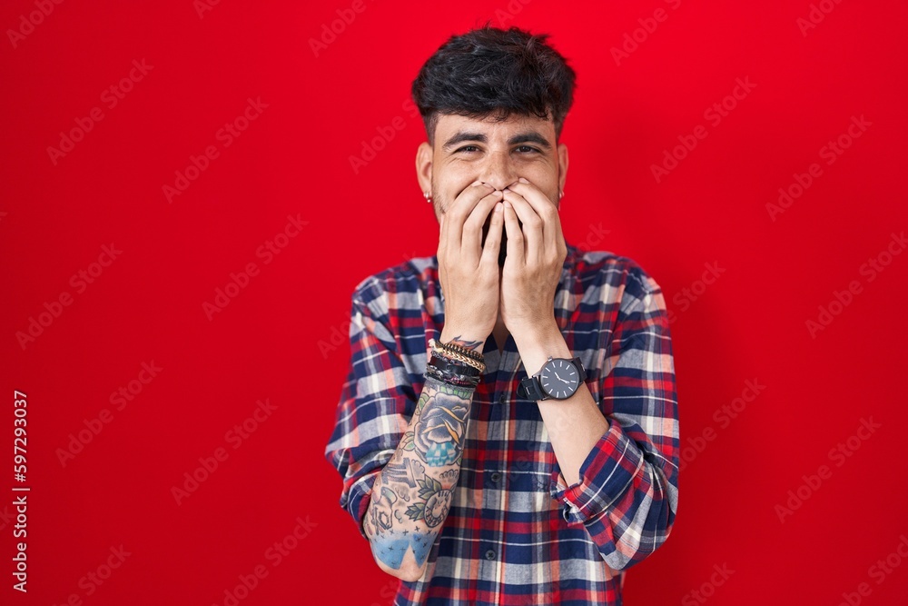 Young hispanic man with beard standing over red background laughing and embarrassed giggle covering mouth with hands, gossip and scandal concept Young hispanic man with beard standing over red background laughing and embarrassed giggle covering mouth with hands, gossip and scandal concept