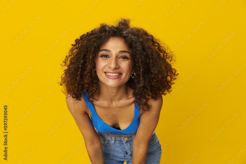 Woman with curly afro hair in a blue t-shirt on. yellow background signs with her hands, look into the camera, smile with teeth and happiness, copy space Woman with curly afro hair in a blue t-shirt on. yellow background signs with her hands, look into the camera, smile with teeth and happiness, copy space