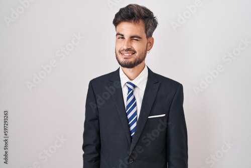 Young hispanic man with tattoos wearing business suit and tie winking looking at the camera with sexy expression, cheerful and happy face.