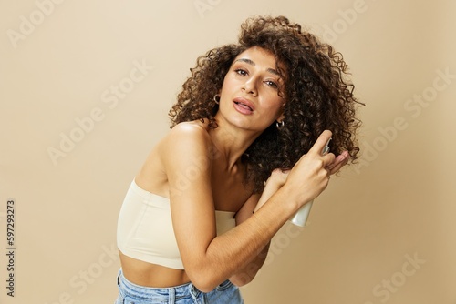 Woman applies cream and balm to her curly hair, the concept of protection and care with salon products, a healthy look, a smile with teeth on a beige background