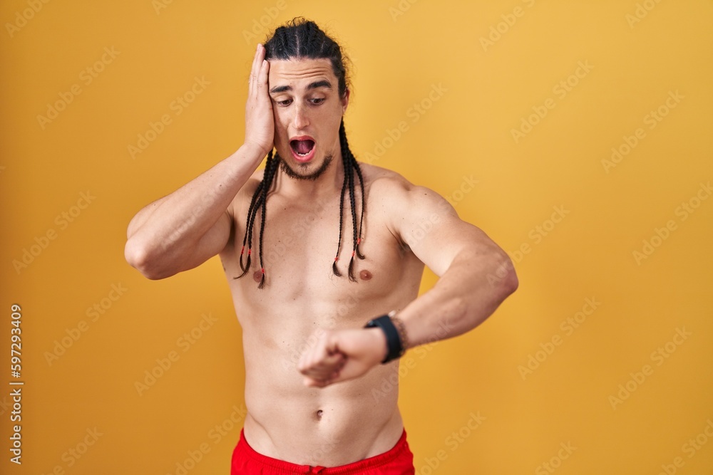 Fototapeta premium Hispanic man with long hair standing shirtless over yellow background looking at the watch time worried, afraid of getting late