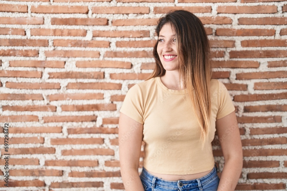 Young brunette woman standing over bricks wall looking away to side with smile on face, natural expression. laughing confident.