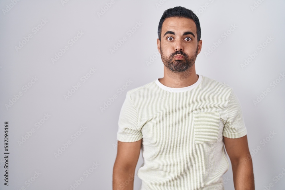 Hispanic man with beard standing over isolated background puffing cheeks with funny face. mouth inflated with air, crazy expression. Hispanic man with beard standing over isolated background puffing cheeks with funny face. mouth inflated with air, crazy expression.