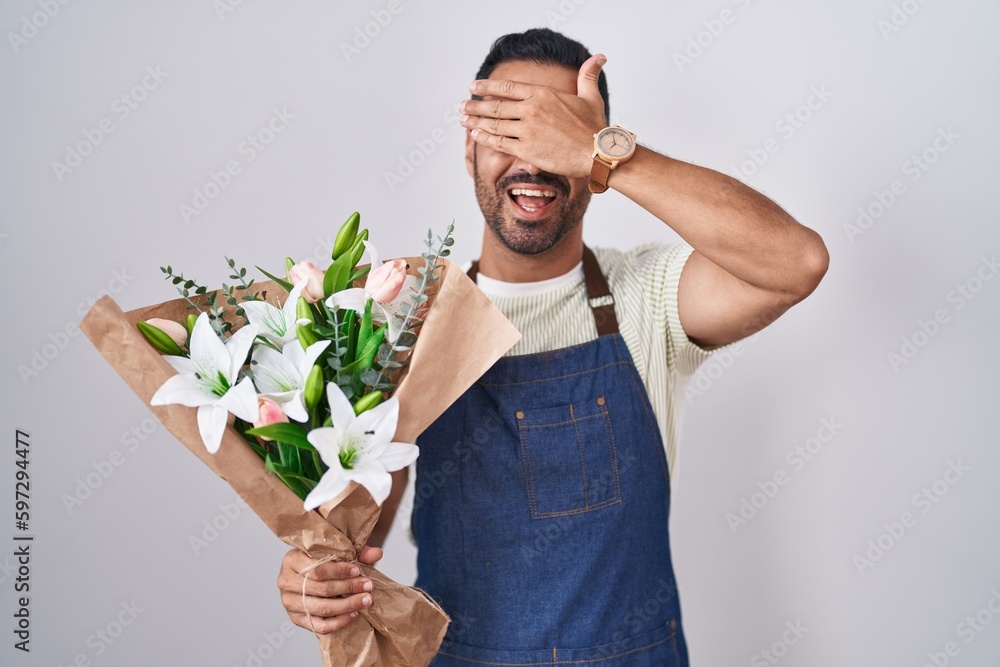 Hispanic man with beard working as florist smiling and laughing with hand on face covering eyes for surprise. blind concept. Hispanic man with beard working as florist smiling and laughing with hand on face covering eyes for surprise. blind concept.