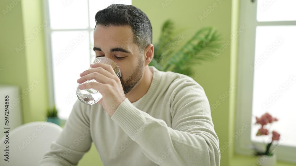 Young hispanic man drinking water sitting on table at home Young hispanic man drinking water sitting on table at home