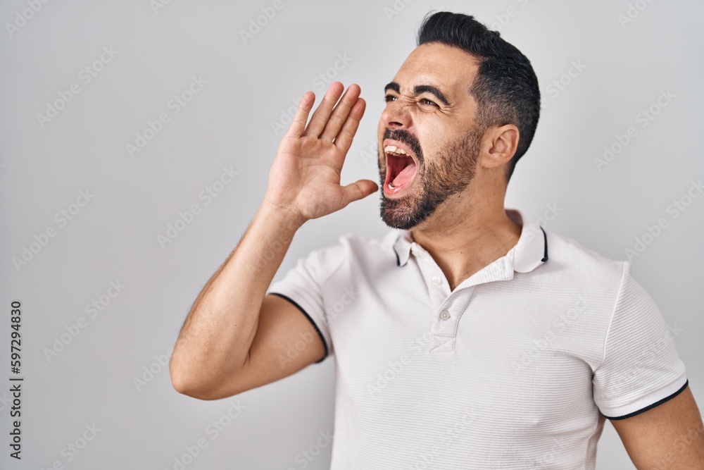 Young hispanic man with beard wearing casual clothes over white background shouting and screaming loud to side with hand on mouth. communication concept. Young hispanic man with beard wearing casual clothes over white background shouting and screaming loud to side with hand on mouth. communication concept.