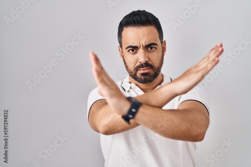 Young hispanic man with beard wearing casual clothes over white background rejection expression crossing arms doing negative sign, angry face