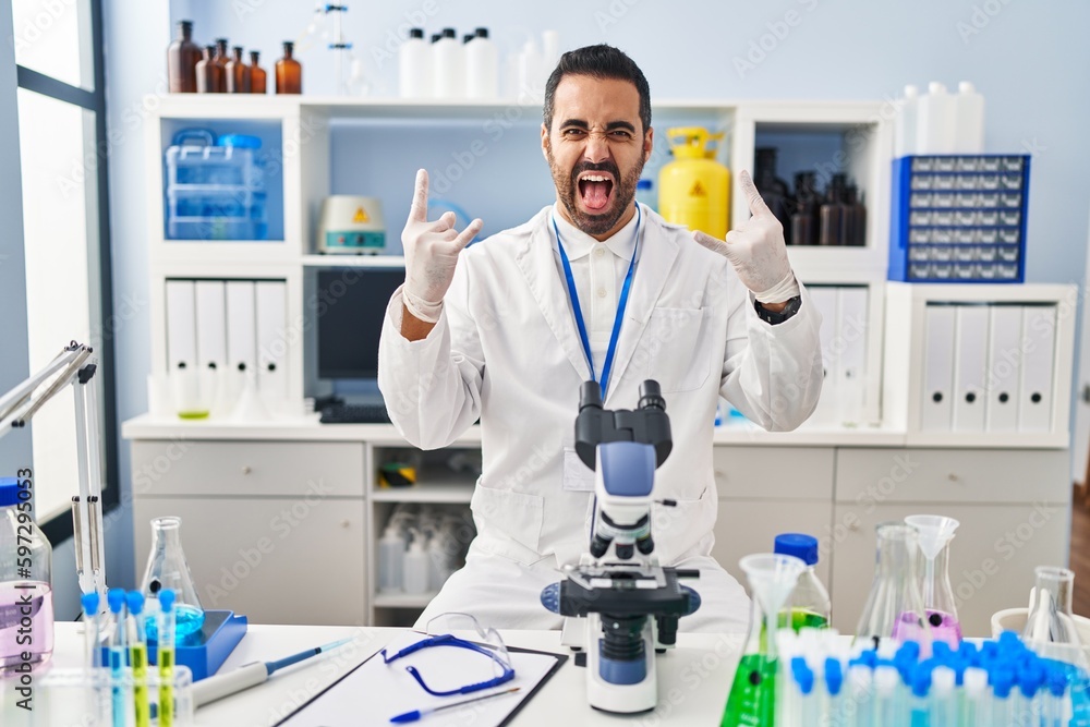Young hispanic man with beard working at scientist laboratory shouting with crazy expression doing rock symbol with hands up. music star. heavy concept. Young hispanic man with beard working at scientist laboratory shouting with crazy expression doing rock symbol with hands up. music star. heavy concept.
