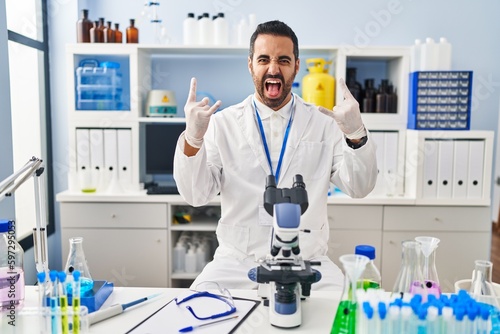 Young hispanic man with beard working at scientist laboratory shouting with crazy expression doing rock symbol with hands up. music star. heavy concept.