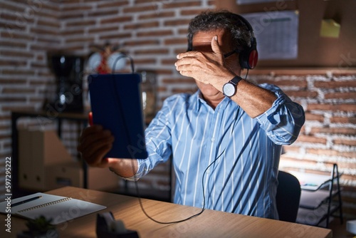 Middle age hispanic man using touchpad sitting on the table at night smiling and laughing with hand on face covering eyes for surprise. blind concept.