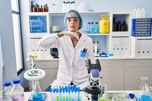Brunette woman working at scientist laboratory cutting throat with hand as knife, threaten aggression with furious violence