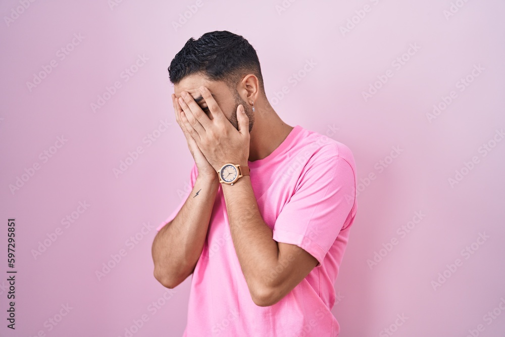 Hispanic young man standing over pink background with sad expression covering face with hands while crying. depression concept. Hispanic young man standing over pink background with sad expression covering face with hands while crying. depression concept.