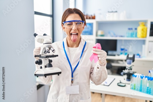 Young brunette woman working at scientist laboratory holding pink ribbon sticking tongue out happy with funny expression.
