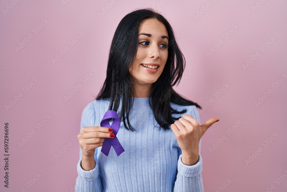 Hispanic woman holding purple ribbon awareness pointing thumb up to the side smiling happy with open mouth Hispanic woman holding purple ribbon awareness pointing thumb up to the side smiling happy with open mouth