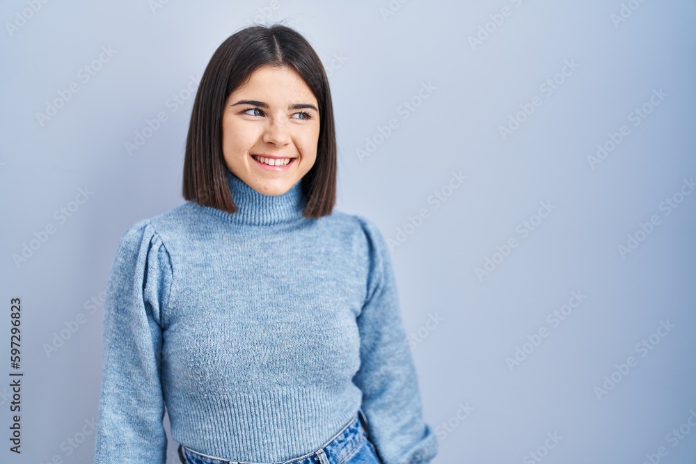 Young hispanic woman standing over blue background looking away to side with smile on face, natural expression. laughing confident. Young hispanic woman standing over blue background looking away to side with smile on face, natural expression. laughing confident.