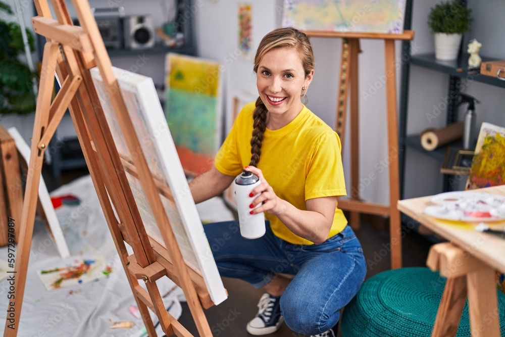 Young blonde woman artist smiling confident using graffiti spray drawing at art studio