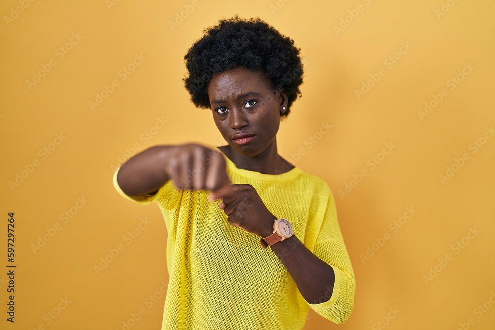 African young woman standing over yellow studio punching fist to fight, aggressive and angry attack, threat and violence African young woman standing over yellow studio punching fist to fight, aggressive and angry attack, threat and violence