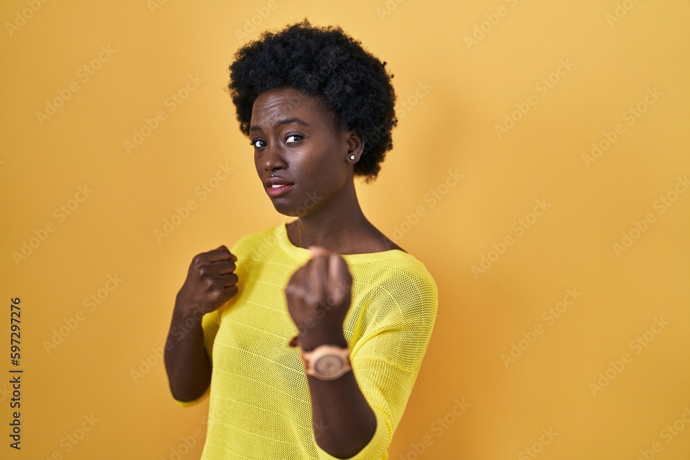 African young woman standing over yellow studio ready to fight with fist defense gesture, angry and upset face, afraid of problem