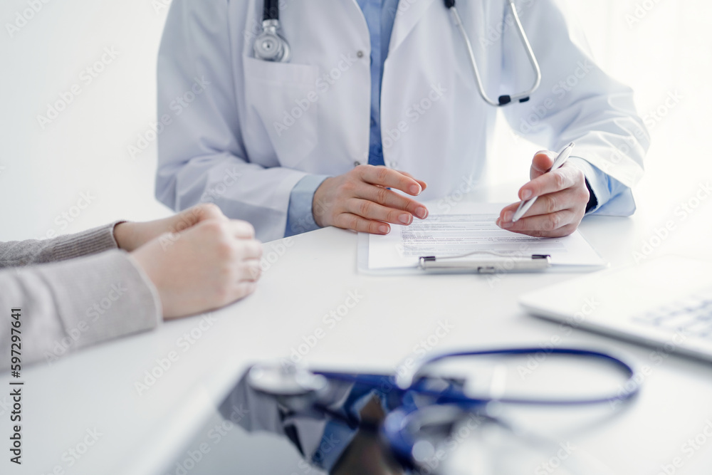 Doctor and patient sitting at the table in clinic. The focus is on female physician's hands filling up the medication history record form or checklist, close up. Medicine concept. Doctor and patient sitting at the table in clinic. The focus is on female physician's hands filling up the medication history record form or checklist, close up. Medicine concept.