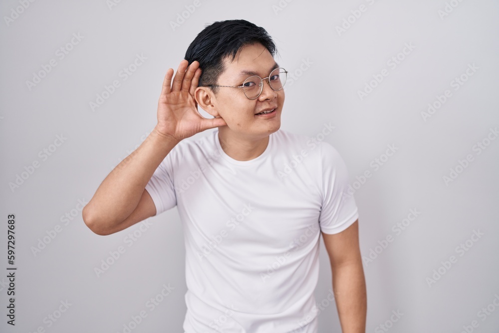 Young asian man standing over white background smiling with hand over ear listening an hearing to rumor or gossip. deafness concept. Young asian man standing over white background smiling with hand over ear listening an hearing to rumor or gossip. deafness concept.