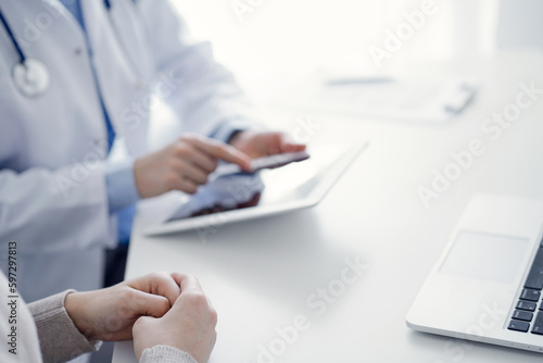 Doctor and patient sitting at the table in clinic while using tablet computer. The focus is on female patient's hands, close up. Medicine concept.