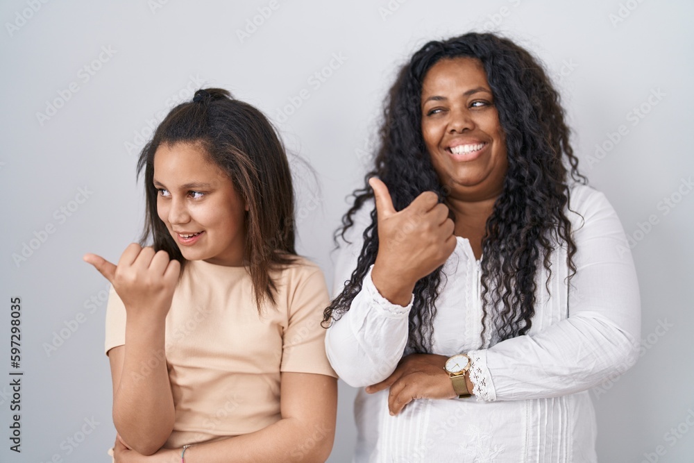 Mother and young daughter standing over white background smiling with happy face looking and pointing to the side with thumb up. Mother and young daughter standing over white background smiling with happy face looking and pointing to the side with thumb up.