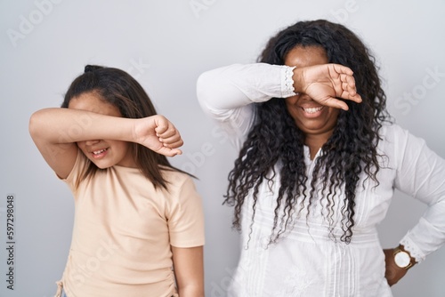 Mother and young daughter standing over white background covering eyes with arm smiling cheerful and funny. blind concept.