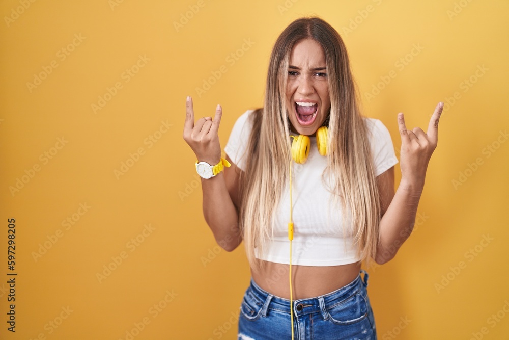Young blonde woman standing over yellow background wearing headphones shouting with crazy expression doing rock symbol with hands up. music star. heavy music concept. Young blonde woman standing over yellow background wearing headphones shouting with crazy expression doing rock symbol with hands up. music star. heavy music concept.