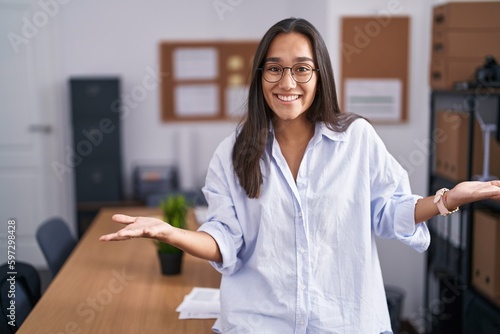 Young hispanic woman at the office smiling cheerful with open arms as friendly welcome, positive and confident greetings © Krakenimages.com