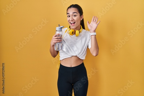 Young south asian woman wearing sportswear drinking water waiving saying hello happy and smiling, friendly welcome gesture