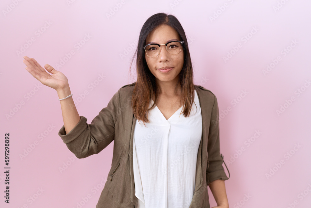 Middle age chinese woman wearing glasses over pink background smiling cheerful presenting and pointing with palm of hand looking at the camera. Middle age chinese woman wearing glasses over pink background smiling cheerful presenting and pointing with palm of hand looking at the camera.
