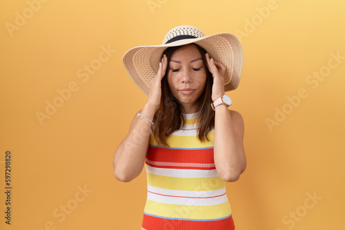 Middle age chinese woman wearing summer hat over yellow background with hand on head, headache because stress. suffering migraine.