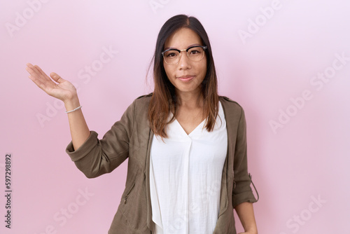 Middle age chinese woman wearing glasses over pink background smiling cheerful presenting and pointing with palm of hand looking at the camera.