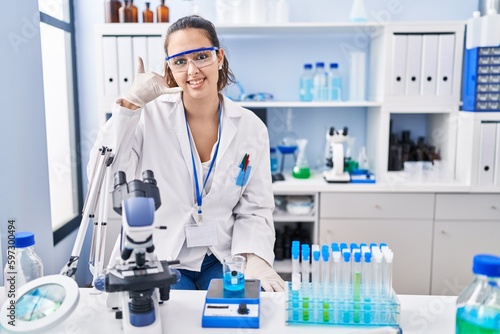 Young hispanic woman working at scientist laboratory smiling doing phone gesture with hand and fingers like talking on the telephone. communicating concepts.