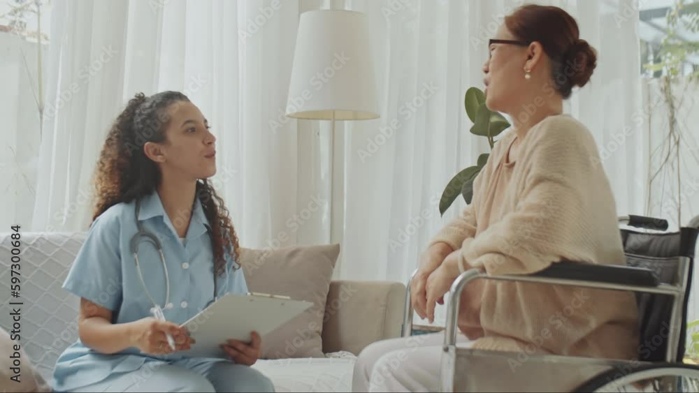 Medium full shot of senior Asian woman in wheelchair explaining symptoms to female doctor during health consultation at home Medium full shot of senior Asian woman in wheelchair explaining symptoms to female doctor during health consultation at home